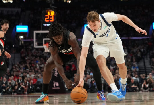 Cooper Flagg #32 of the Dallas Mavericks and Jrue Holiday #5 of the Portland Trail Blazers dive for a loose ball at Moda Center.