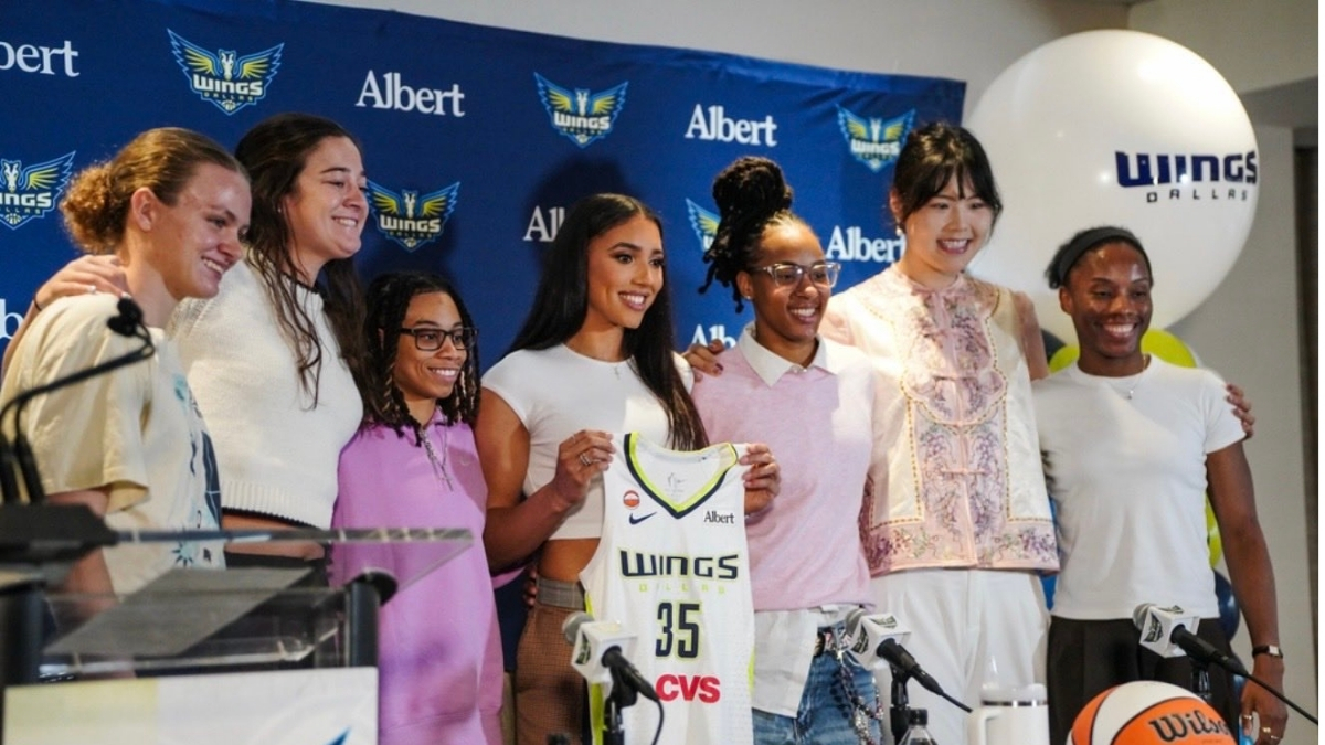 Azzi Fudd holds up her new #35 Dallas Wings jersey while posing with teammates Maddy Siegrist, JJ Quinerly, Grace Berger, Aziaha James, Li Yueru, and Shyanne Sellers at her introductory press conference.