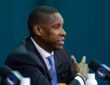 Profile view of Masai Ujiri gesturing while speaking into a microphone at a podium featuring the Dallas Mavericks logo.