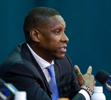 Profile view of Masai Ujiri gesturing while speaking into a microphone at a podium featuring the Dallas Mavericks logo.