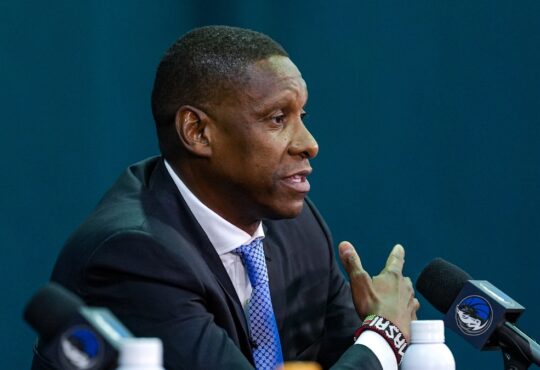 Profile view of Masai Ujiri gesturing while speaking into a microphone at a podium featuring the Dallas Mavericks logo.