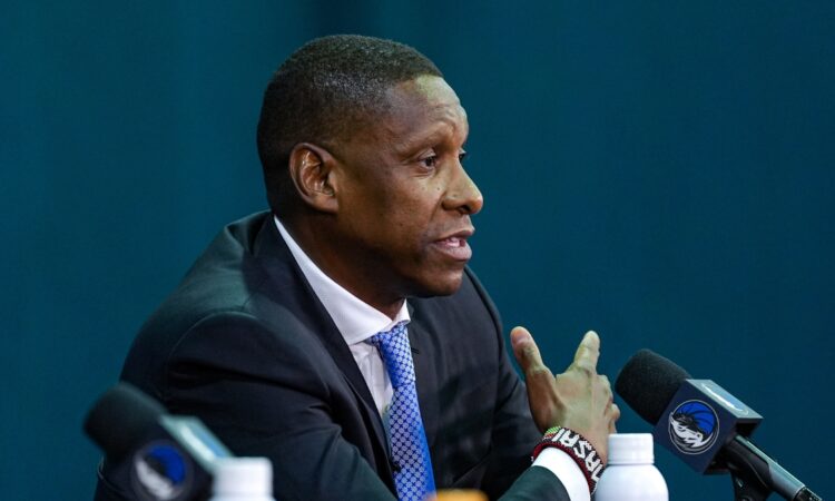 Profile view of Masai Ujiri gesturing while speaking into a microphone at a podium featuring the Dallas Mavericks logo.