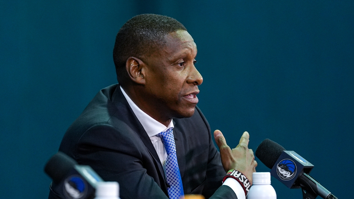 Profile view of Masai Ujiri gesturing while speaking into a microphone at a podium featuring the Dallas Mavericks logo.