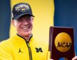 Dusty May smiles while holding the 2026 NCAA national championship trophy in a Michigan quarter-zip and Champions cap