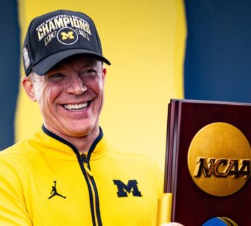Dusty May smiles while holding the 2026 NCAA national championship trophy in a Michigan quarter-zip and Champions cap