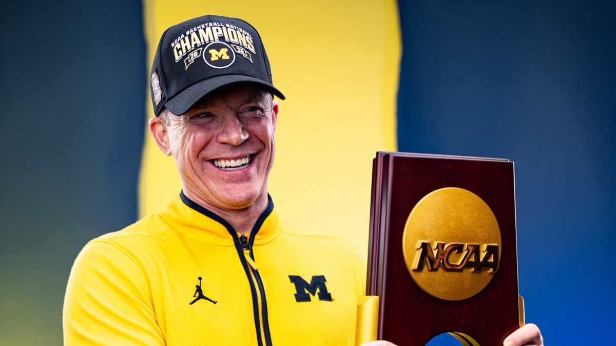 Dusty May smiles while holding the 2026 NCAA national championship trophy in a Michigan quarter-zip and Champions cap