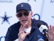 Dallas Cowboys owner Jerry Jones, in a navy Cowboys cap and sunglasses, raises both hands while speaking at a podium microphone during the team's training camp opening press conference in Oxnard, California.