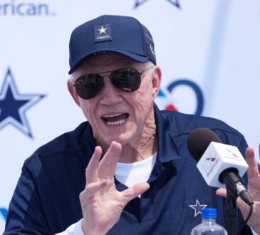 Dallas Cowboys owner Jerry Jones, in a navy Cowboys cap and sunglasses, raises both hands while speaking at a podium microphone during the team's training camp opening press conference in Oxnard, California.