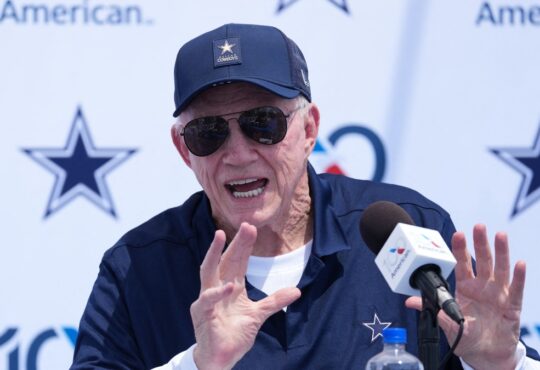 Dallas Cowboys owner Jerry Jones, in a navy Cowboys cap and sunglasses, raises both hands while speaking at a podium microphone during the team's training camp opening press conference in Oxnard, California.
