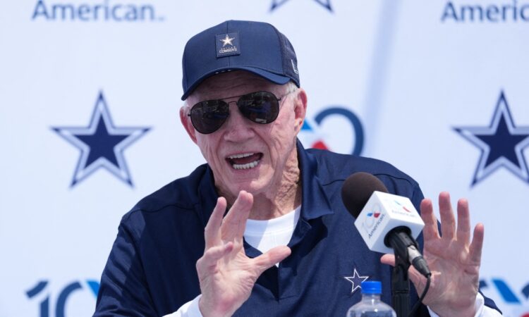 Dallas Cowboys owner Jerry Jones, in a navy Cowboys cap and sunglasses, raises both hands while speaking at a podium microphone during the team's training camp opening press conference in Oxnard, California.