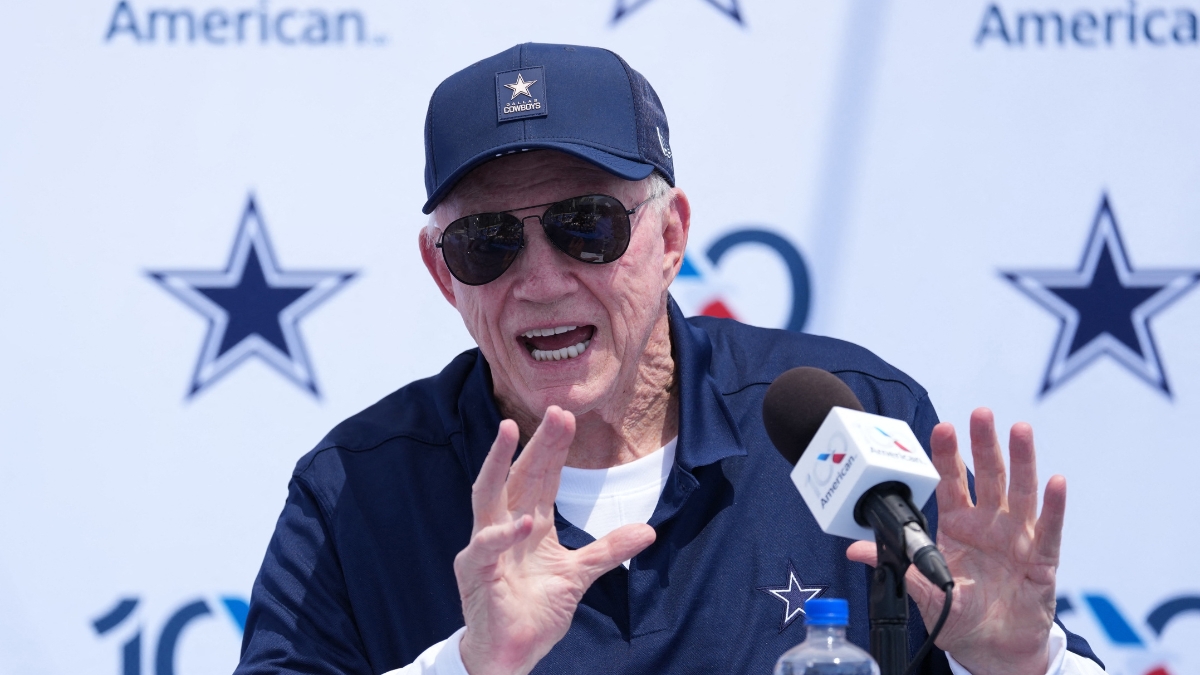 Dallas Cowboys owner Jerry Jones, in a navy Cowboys cap and sunglasses, raises both hands while speaking at a podium microphone during the team's training camp opening press conference in Oxnard, California.