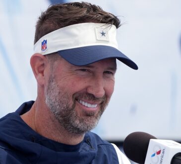 Dallas Cowboys head coach Brian Schottenheimer smiles while speaking at a microphone during a press conference.