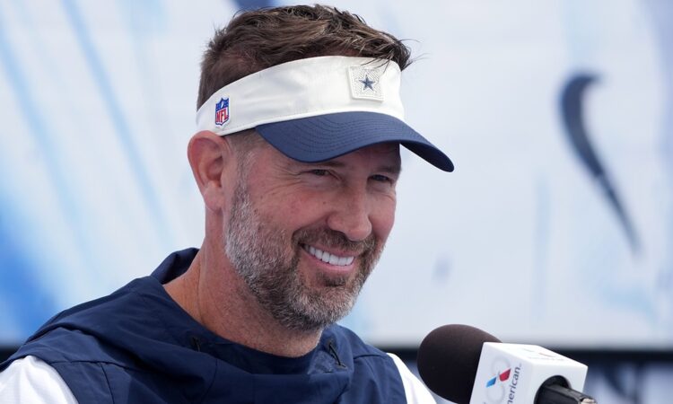 Dallas Cowboys head coach Brian Schottenheimer smiles while speaking at a microphone during a press conference.