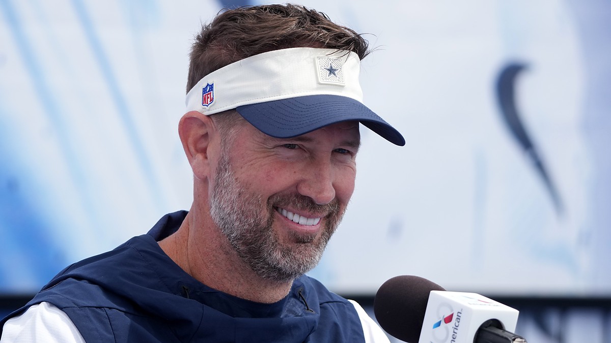 Dallas Cowboys head coach Brian Schottenheimer smiles while speaking at a microphone during a press conference.