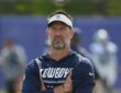 Dallas Cowboys head coach Brian Schottenheimer stands with his arms crossed, watching practice in a Cowboys visor and hoodie.