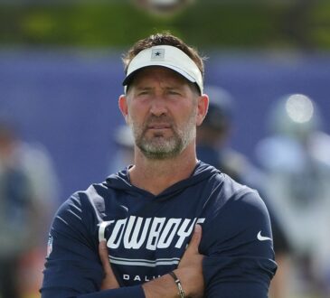 Dallas Cowboys head coach Brian Schottenheimer stands with his arms crossed, watching practice in a Cowboys visor and hoodie.