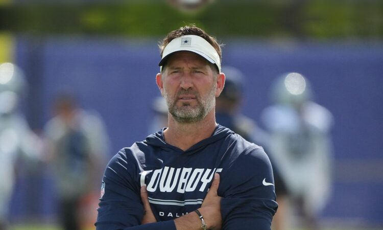 Dallas Cowboys head coach Brian Schottenheimer stands with his arms crossed, watching practice in a Cowboys visor and hoodie.