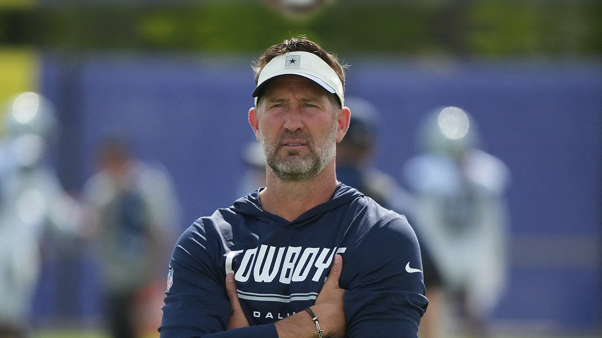 Dallas Cowboys head coach Brian Schottenheimer stands with his arms crossed, watching practice in a Cowboys visor and hoodie.