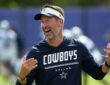 Dallas Cowboys head coach Brian Schottenheimer laughs with both hands raised on the practice field.