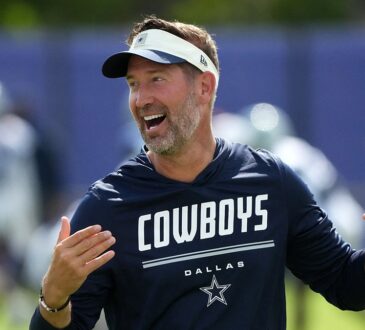 Dallas Cowboys head coach Brian Schottenheimer laughs with both hands raised on the practice field.
