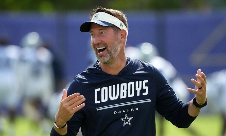Dallas Cowboys head coach Brian Schottenheimer laughs with both hands raised on the practice field.