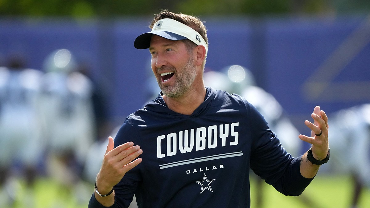 Dallas Cowboys head coach Brian Schottenheimer laughs with both hands raised on the practice field.
