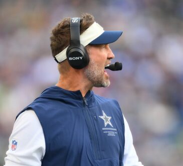 Dallas Cowboys head coach Brian Schottenheimer calls out from the sideline wearing a headset.