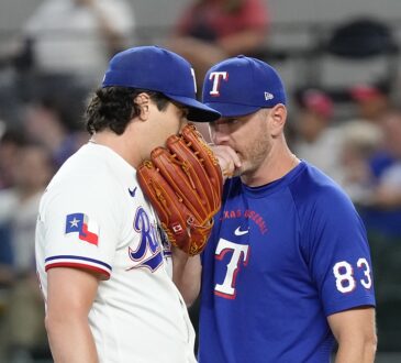 Texas Rangers pitching coach Jordan Tiegs talks with pitcher Cal Quantrill on the mound, both covering their mouths.