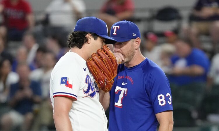 Texas Rangers pitching coach Jordan Tiegs talks with pitcher Cal Quantrill on the mound, both covering their mouths.