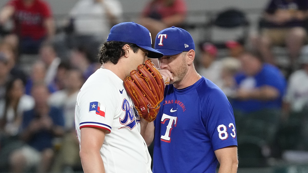 Texas Rangers pitching coach Jordan Tiegs talks with pitcher Cal Quantrill on the mound, both covering their mouths.