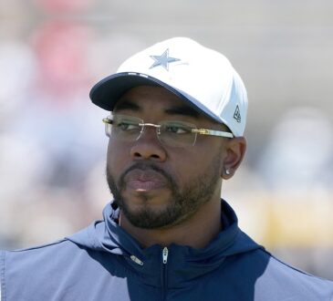 Dallas Cowboys defensive coordinator Christian Parker watches practice in a white Cowboys cap and navy vest.
