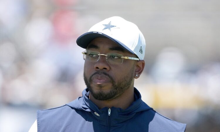 Dallas Cowboys defensive coordinator Christian Parker watches practice in a white Cowboys cap and navy vest.