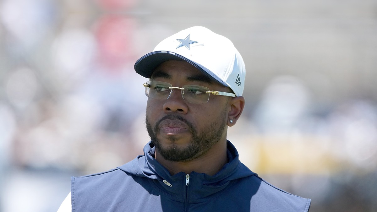 Dallas Cowboys defensive coordinator Christian Parker watches practice in a white Cowboys cap and navy vest.