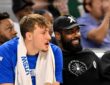 Dallas Mavericks forward Cooper Flagg and guard Kyrie Irving sit together on the bench, Irving smiling in a black cap and hoodie.