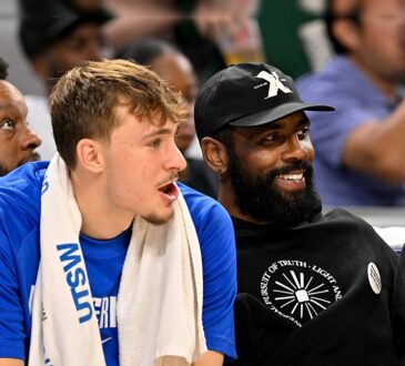Dallas Mavericks forward Cooper Flagg and guard Kyrie Irving sit together on the bench, Irving smiling in a black cap and hoodie.