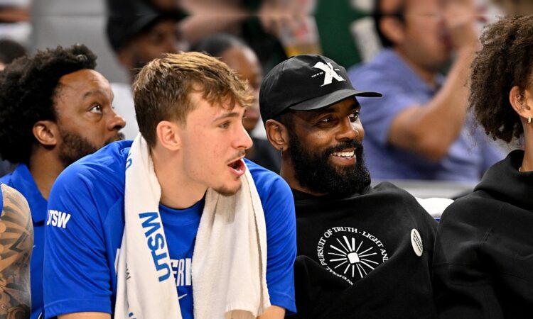 Dallas Mavericks forward Cooper Flagg and guard Kyrie Irving sit together on the bench, Irving smiling in a black cap and hoodie.