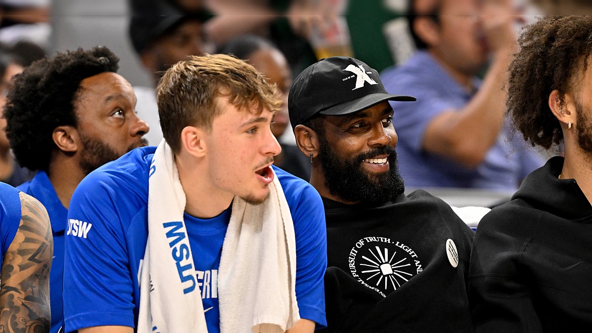 Dallas Mavericks forward Cooper Flagg and guard Kyrie Irving sit together on the bench, Irving smiling in a black cap and hoodie.