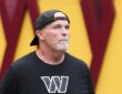 Washington Commanders head coach Dan Quinn looks up as he walks onto the field in a backward Commanders cap.