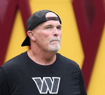 Washington Commanders head coach Dan Quinn looks up as he walks onto the field in a backward Commanders cap.