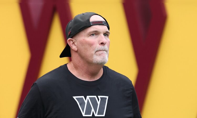 Washington Commanders head coach Dan Quinn looks up as he walks onto the field in a backward Commanders cap.