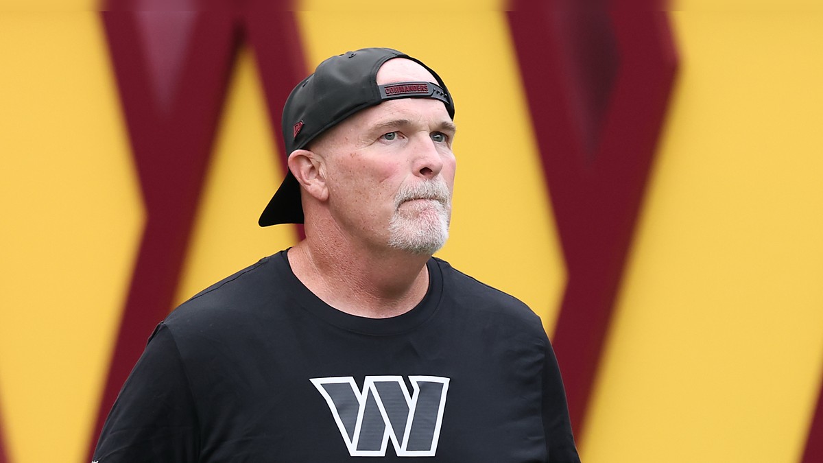 Washington Commanders head coach Dan Quinn looks up as he walks onto the field in a backward Commanders cap.