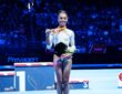 WOGA Plano gymnast Hezly Rivera smiles as she holds up her first-place medal and a bouquet on the competition floor.