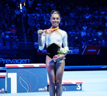 WOGA Plano gymnast Hezly Rivera smiles as she holds up her first-place medal and a bouquet on the competition floor.