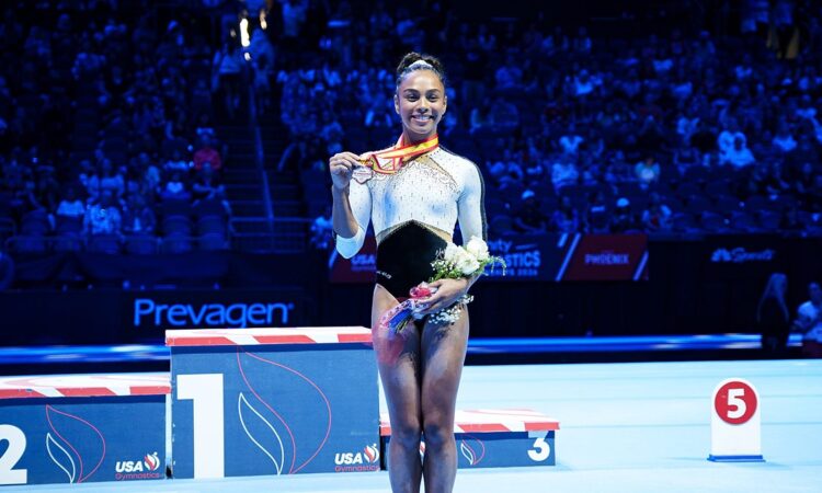 WOGA Plano gymnast Hezly Rivera smiles as she holds up her first-place medal and a bouquet on the competition floor.