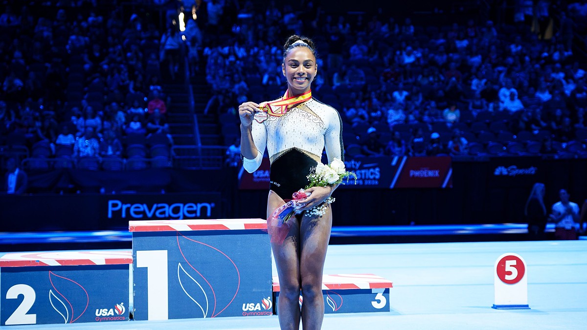 WOGA Plano gymnast Hezly Rivera smiles as she holds up her first-place medal and a bouquet on the competition floor.