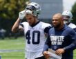 Dallas Cowboys running back Hunter Luepke pulls off his helmet as he walks off the practice field beside a coach.