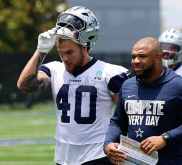 Dallas Cowboys running back Hunter Luepke pulls off his helmet as he walks off the practice field beside a coach.