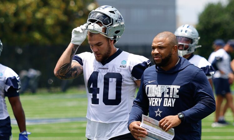 Dallas Cowboys running back Hunter Luepke pulls off his helmet as he walks off the practice field beside a coach.