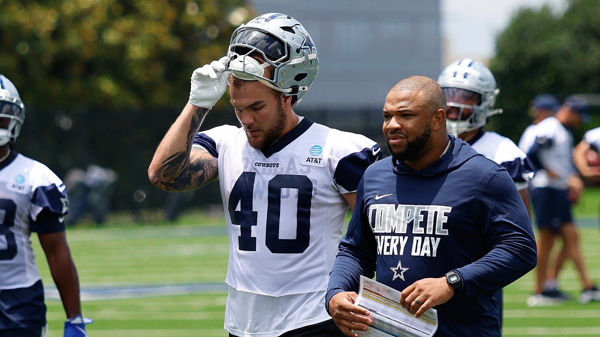 Dallas Cowboys running back Hunter Luepke pulls off his helmet as he walks off the practice field beside a coach.