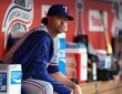 Texas Rangers pitcher Jacob deGrom sits on the dugout rail in a warmup jacket before a game.